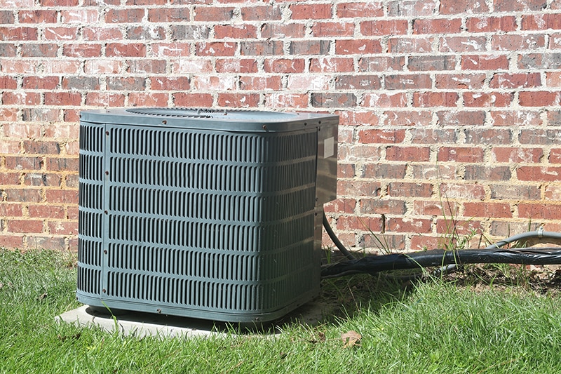 Should You Repair or Replace Your Air Conditioner? Home Air Conditioner Condenser coil sitting in front of brick wall.